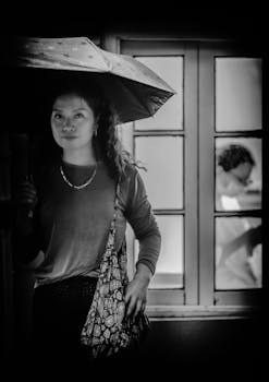 Elegant black and white photo of a woman holding an umbrella, standing by a window.