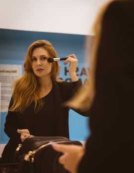 A woman uses a makeup brush in front of a mirror, reflecting her image.