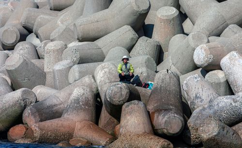 Free A solitary fisherman sits on massive concrete tetrapods by the waterfront, enjoying a calm shore day. Stock Photo
