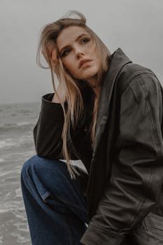 A fashionable woman in a leather jacket poses thoughtfully by the seaside, capturing a moody aesthetic.