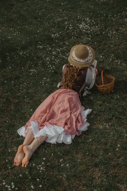 Woman in a pink dress lying barefoot on grass, wearing a straw hat and reading. Peaceful outdoor setting.