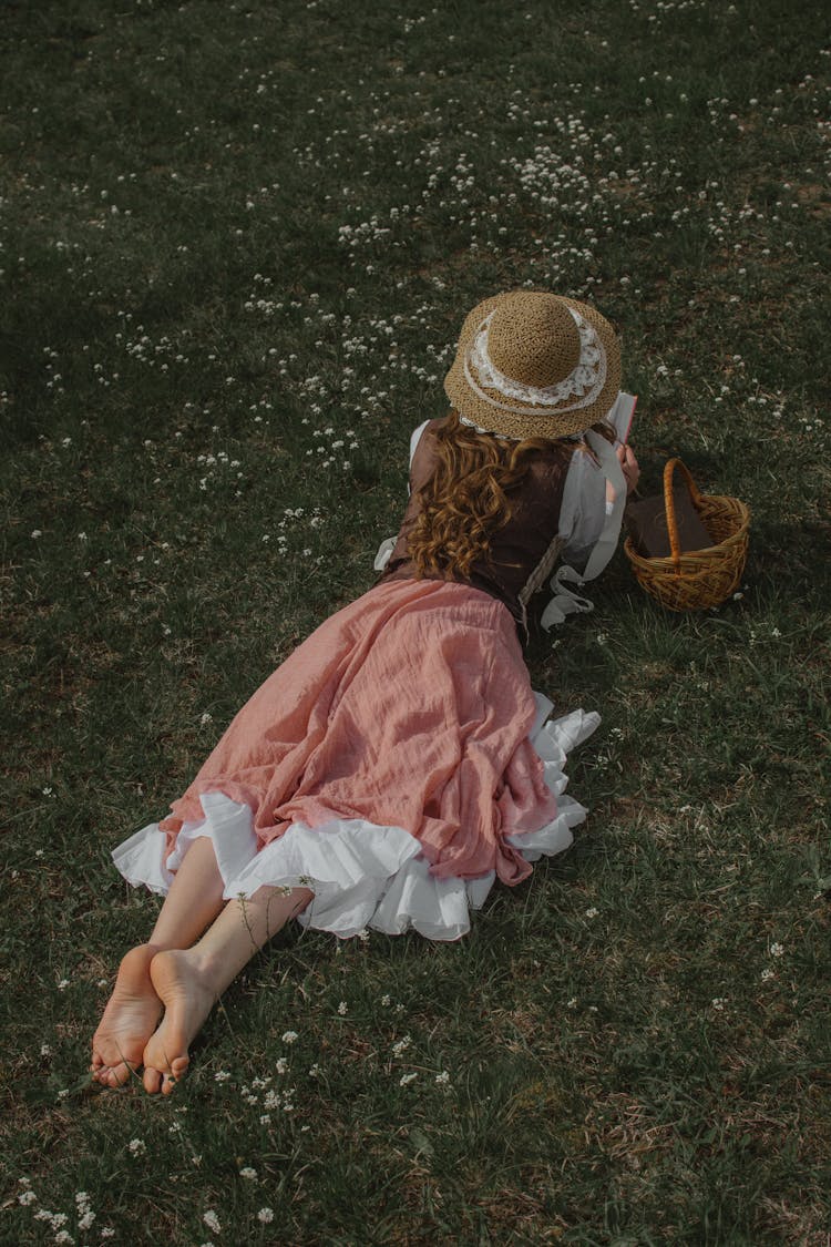 Woman In Pink Dress And Straw Hat Lying On Grass Reading