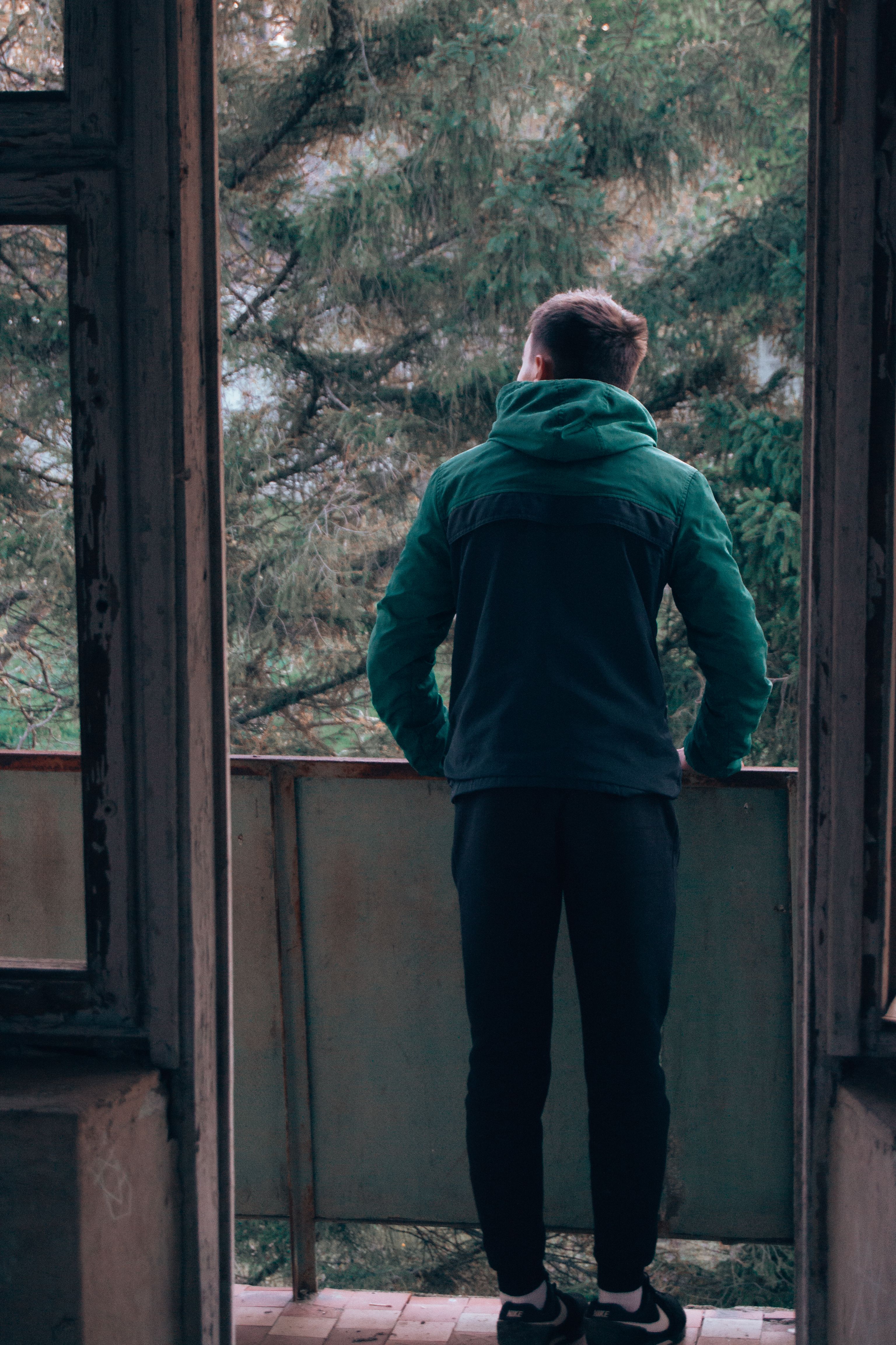 A man in a green jacket stands on a balcony, looking at a forest view.