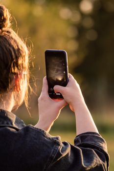 A woman takes a photo of the sunset using her smartphone, featuring a blurred background.
