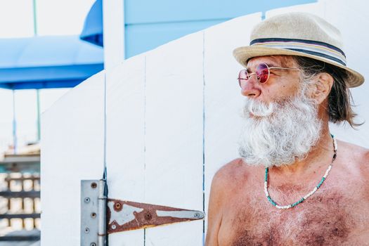 Elderly man with a gray beard and hat enjoying a sunny day by the beach.