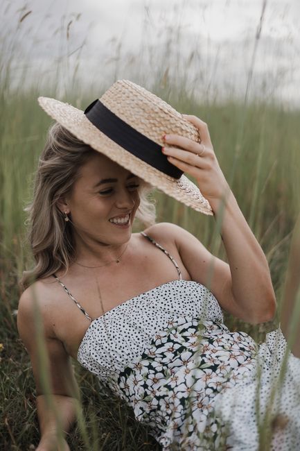 Smiling woman in floral dress holding a straw hat in a grassy field during summer.