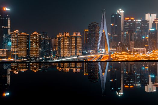 Stunning nighttime view of a modern city skyline reflected in water, showcasing illuminated skyscrapers and a bridge.