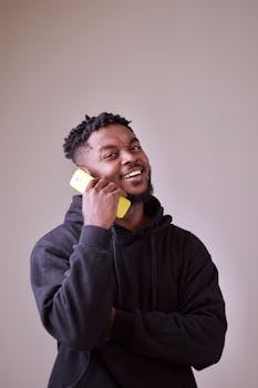 Happy man wearing a black hoodie, talking on a yellow mobile phone indoors, captured in a studio setting.