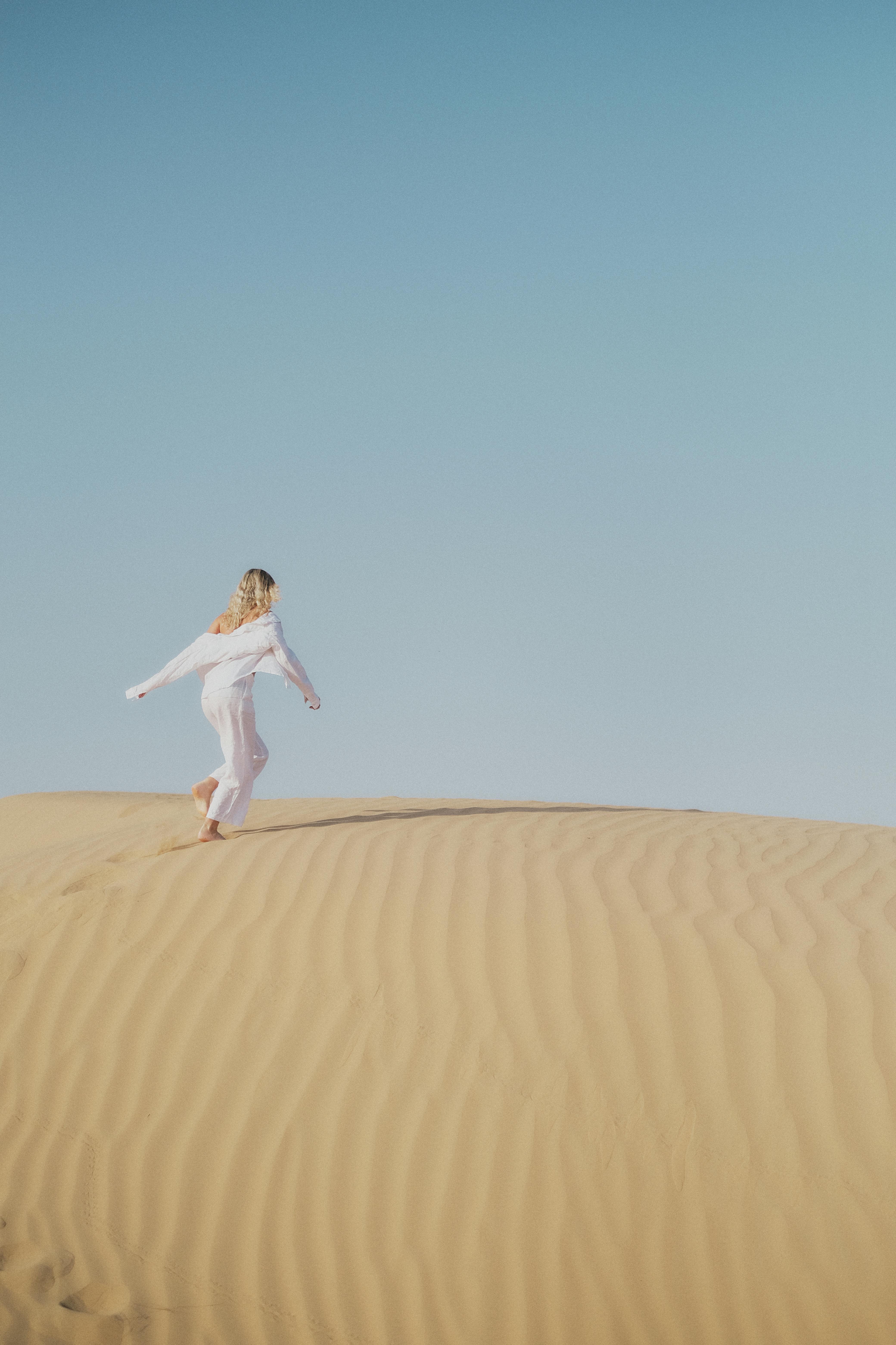A woman in a white dress runs gracefully on the sand dunes under a clear blue sky in Dubai.