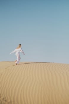 A woman in a white dress runs gracefully on the sand dunes under a clear blue sky in Dubai.