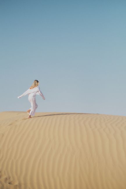 A woman in a white dress runs gracefully on the sand dunes under a clear blue sky in Dubai.