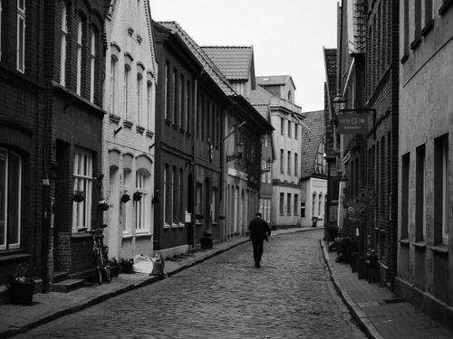 Free Monochrome image of a person walking along a cobblestone street flanked by historical buildings. Stock Photo
