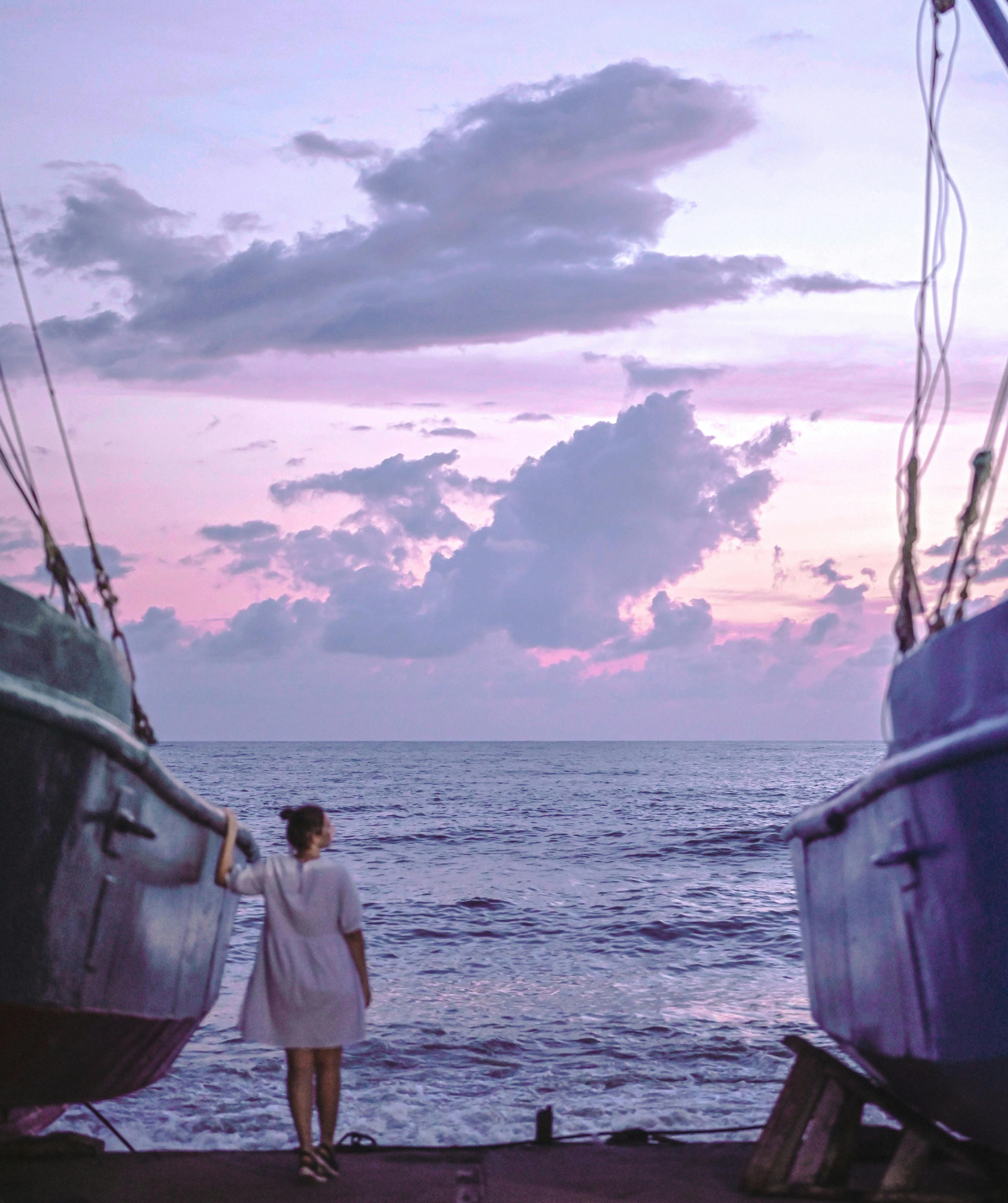 Woman standing between boats, enjoying a pastel sunset over the sea.