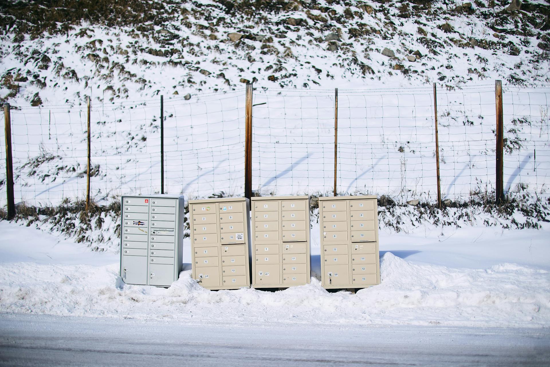 A row of mailboxes partially covered with snow against a fence during winter.