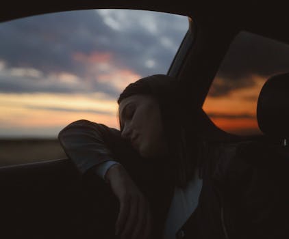 A serene woman rests inside a car, gazing at a vibrant sunset through the window.