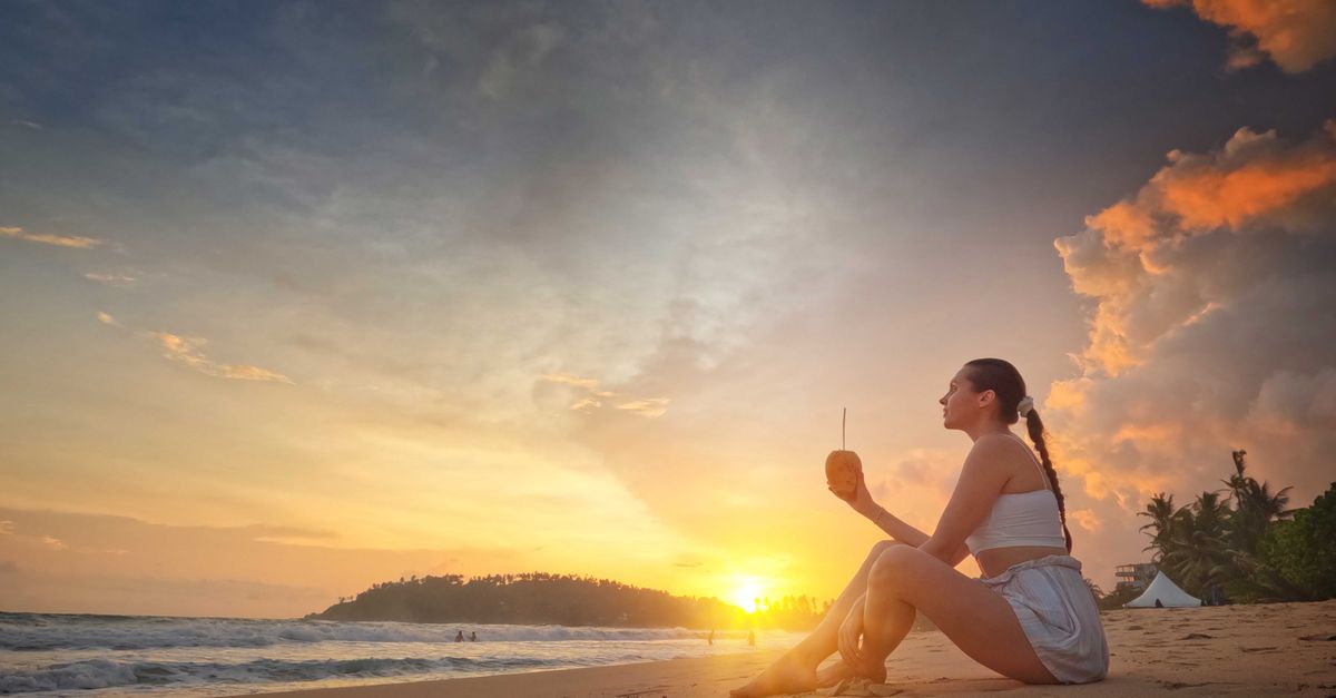 Woman sitting on Mirissa Beach during sunset, enjoying a tropical drink with vibrant sky and waves.