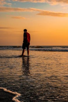 A man walks along a serene beach at sunset, creating a peaceful reflection in the water.