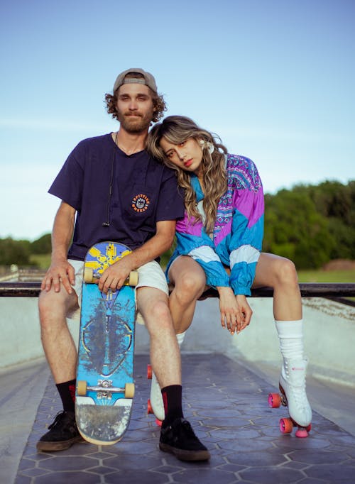 Man Holding a Skateboard and Woman Wearing Roller Skates Sitting in a Skatepark