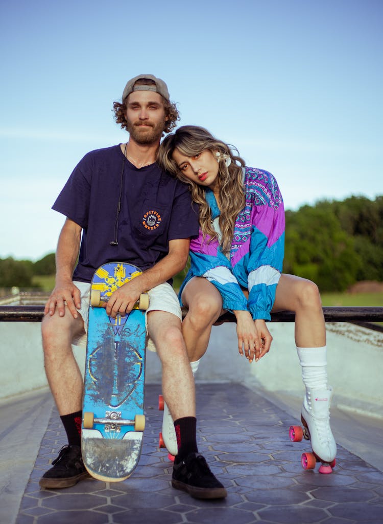 Man Holding A Skateboard And Woman Wearing Roller Skates Sitting In A Skatepark