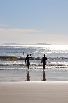 Two surfers walking into the ocean at sunset on a Brazilian beach, capturing a tranquil tropical scene.
