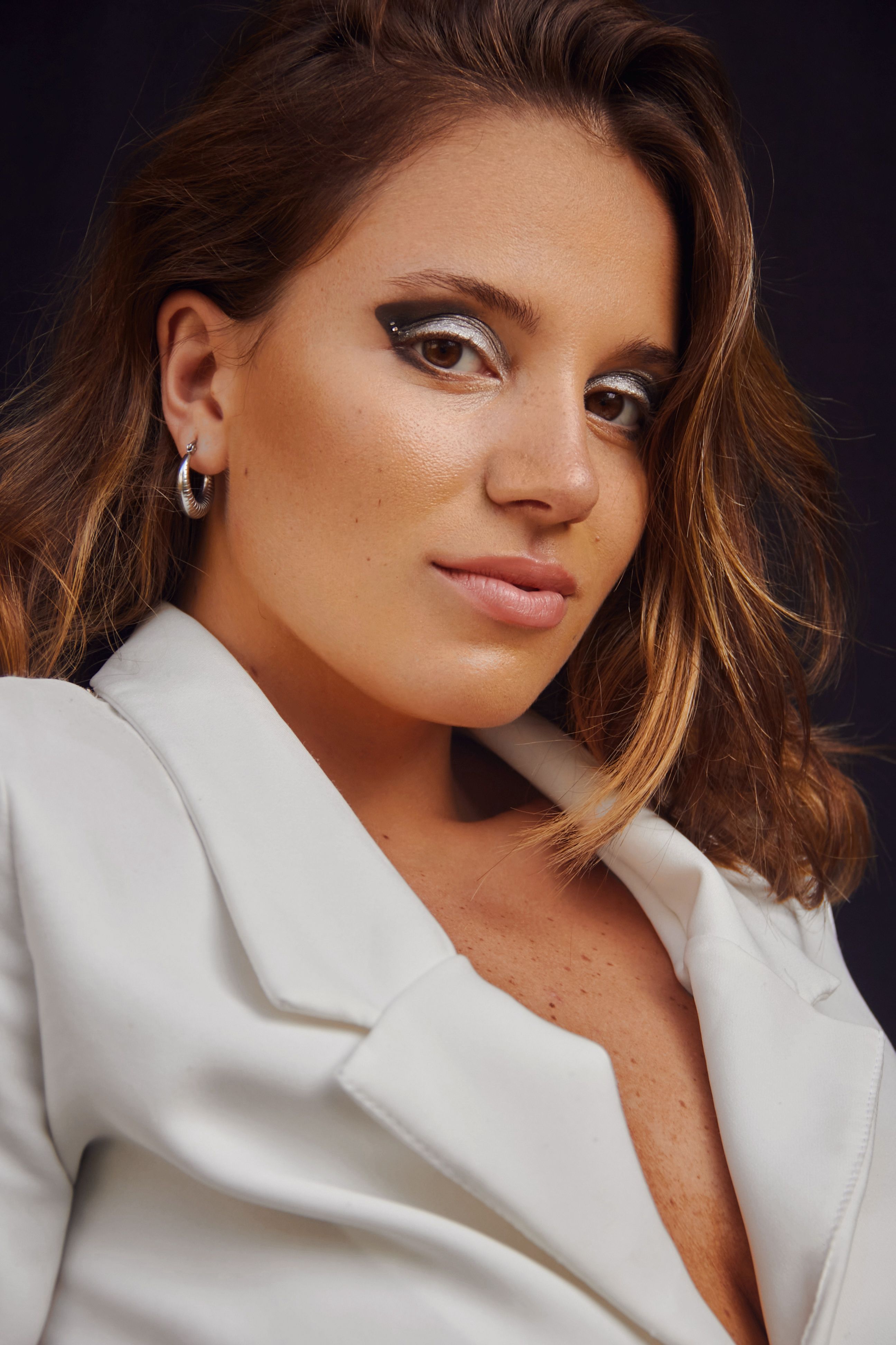 Portrait of a stylish woman in a white blazer with bold makeup against a dark background.