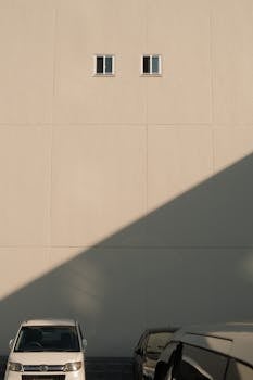 A minimalist building facade with cars and a shadow falling across the wall in daylight.