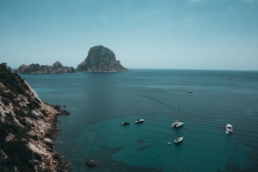 A tranquil view of boats near the rocky coast of Es Vedrà island, Ibiza, under a clear blue sky.