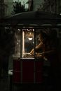 Man in Brown Jacket Sitting on Red and Black Food Stall