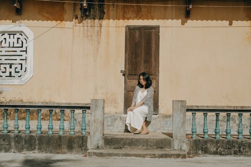 Free Woman in White Dress and Cardigan Sitting In Front of a Wooden Door  Stock Photo