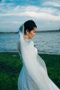 Woman in White Wedding Dress Standing on Green Grass Field Near Body of Water