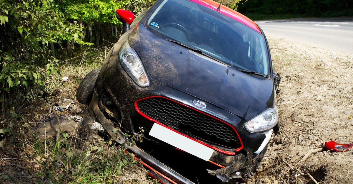 A black car, seemingly a Ford, crashed and damaged in a roadside ditch with surrounding greenery.