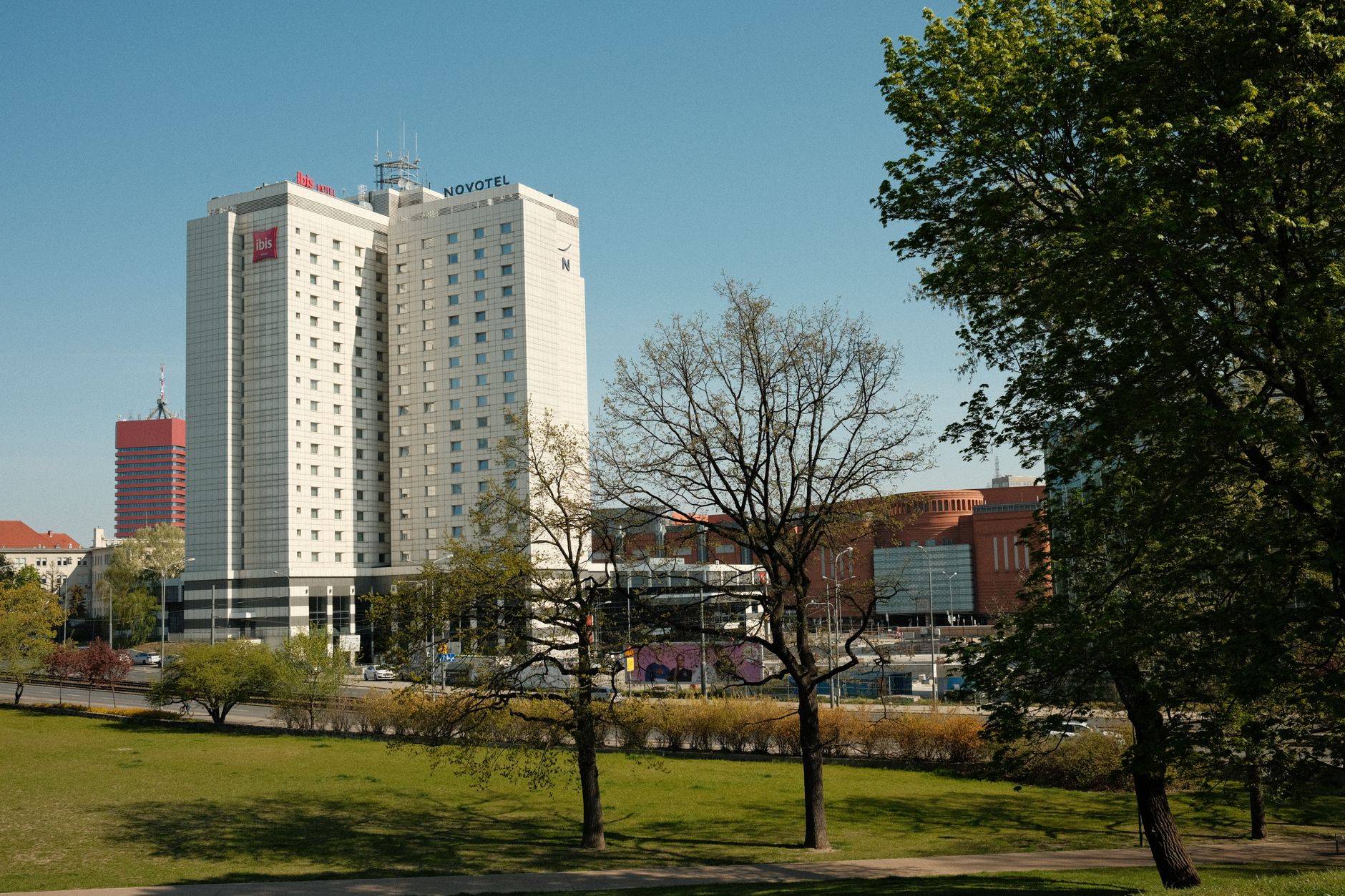 A modern hotel building situated next to a lush urban park under clear blue skies.