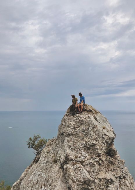 Two people on a rocky cliff gazing at the ocean under a cloudy sky