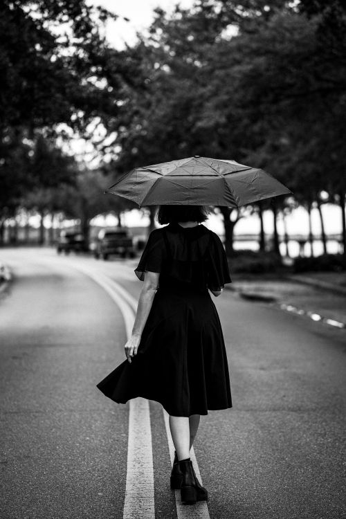 Free A woman with an umbrella strolls down a rain-soaked street in black and white tones. Stock Photo