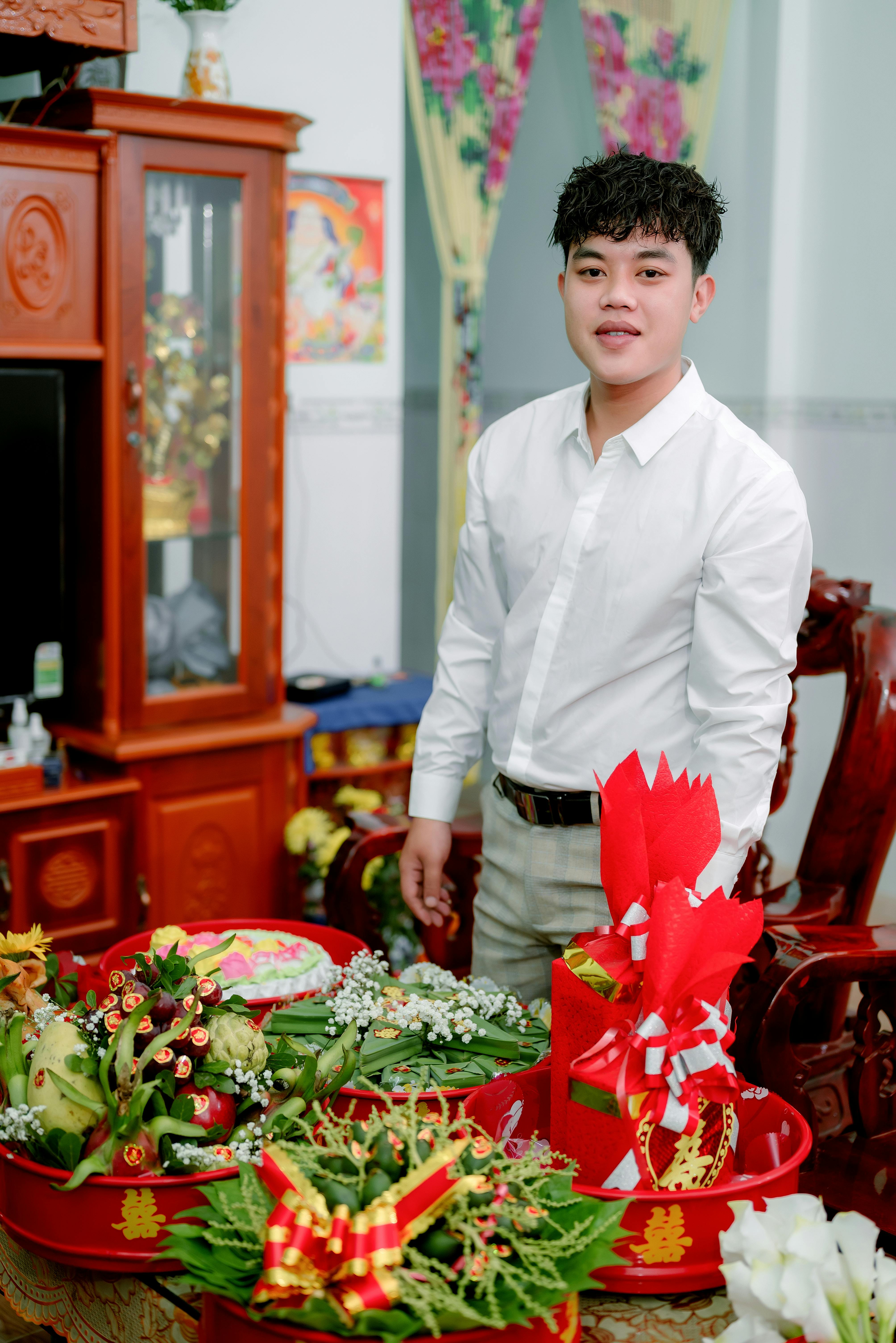 Asian man in white long sleeves standing near festive table indoors, smiling amid floral decorations.