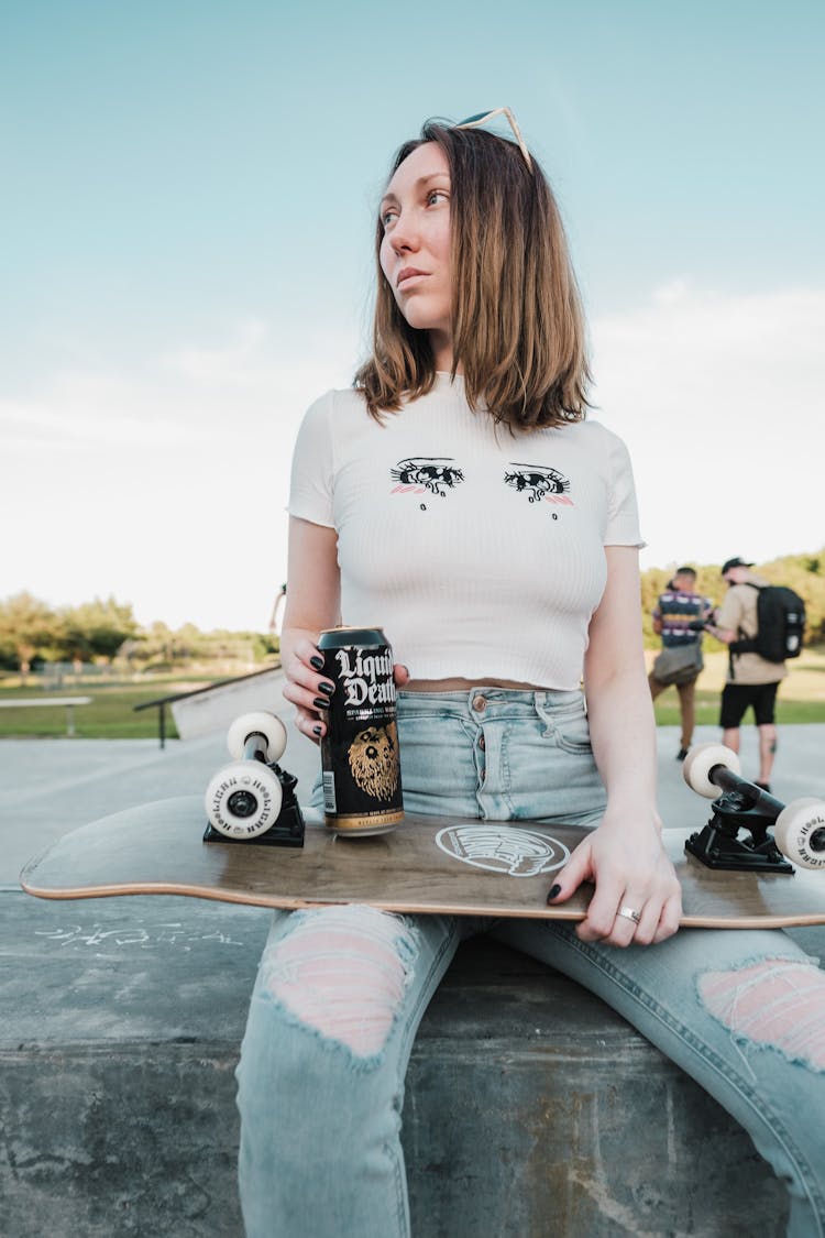 Young Woman Sitting In White Crop Top And Ripped Jeans Holding A Skateboard And Drink In Can