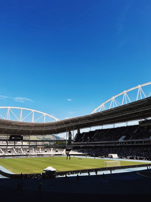 Free Expansive view of a sports stadium filled with spectators under a vivid blue sky, showcasing a soccer field and distinctive architecture. Stock Photo