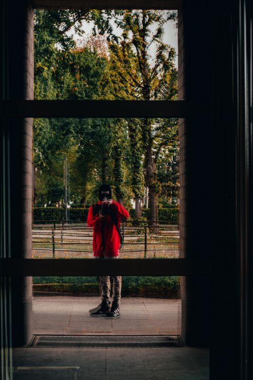 Free Photographer in red jacket reflected in window with trees in background. Stock Photo
