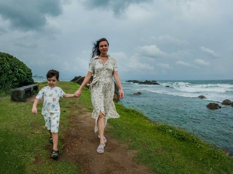 A mother and son enjoy a walk along the scenic coastline of Galle, Sri Lanka under an overcast sky.