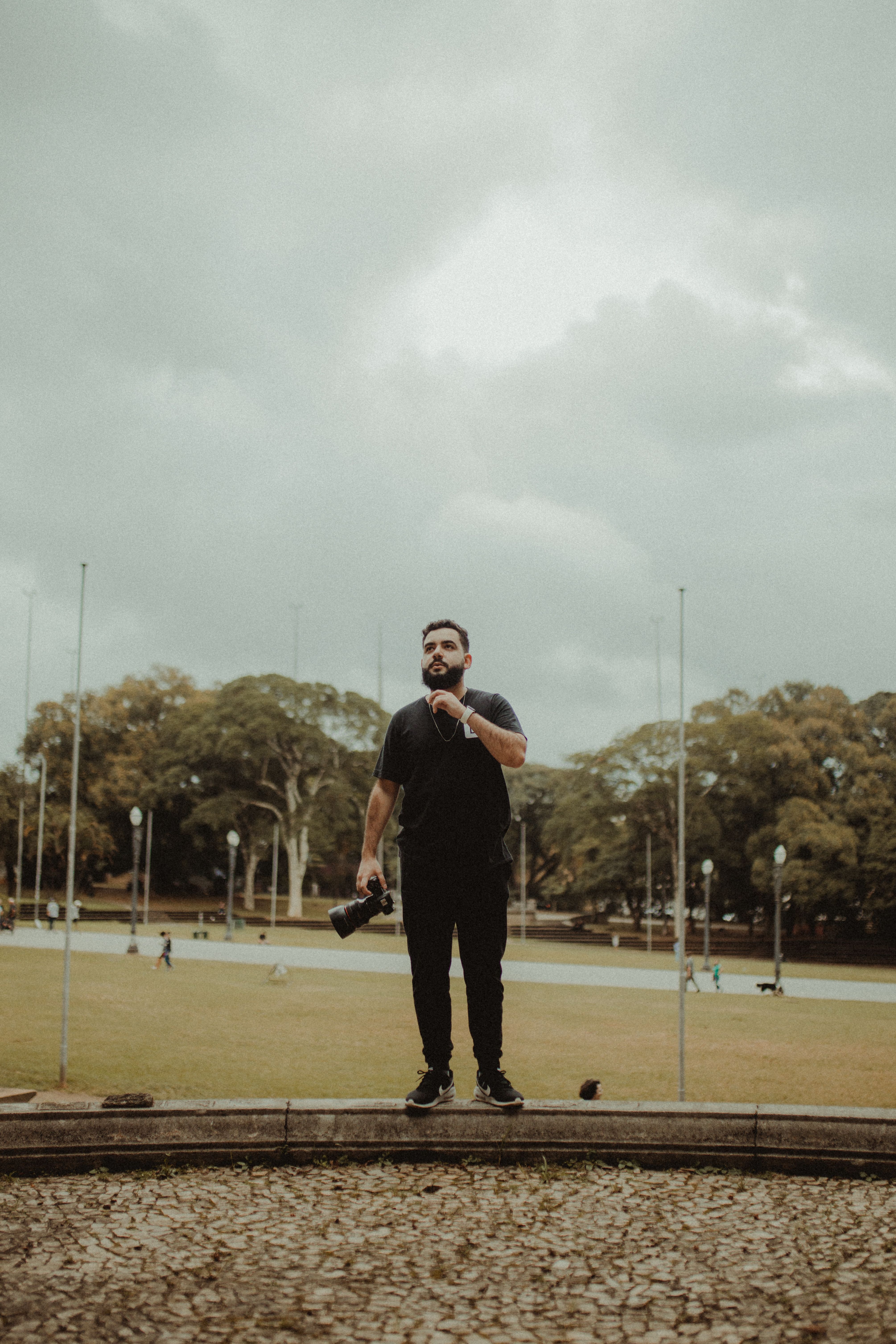 A man holding a camera stands contemplatively in a park under a cloudy sky.