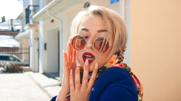 Trendy blond woman with sunglasses and floral scarf posing outdoors, showcasing chic fashion and manicured nails.