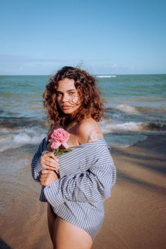 A woman holding a pink rose poses on a sunny beach in Arraial d'Ajuda, Brazil, with ocean waves in the background.