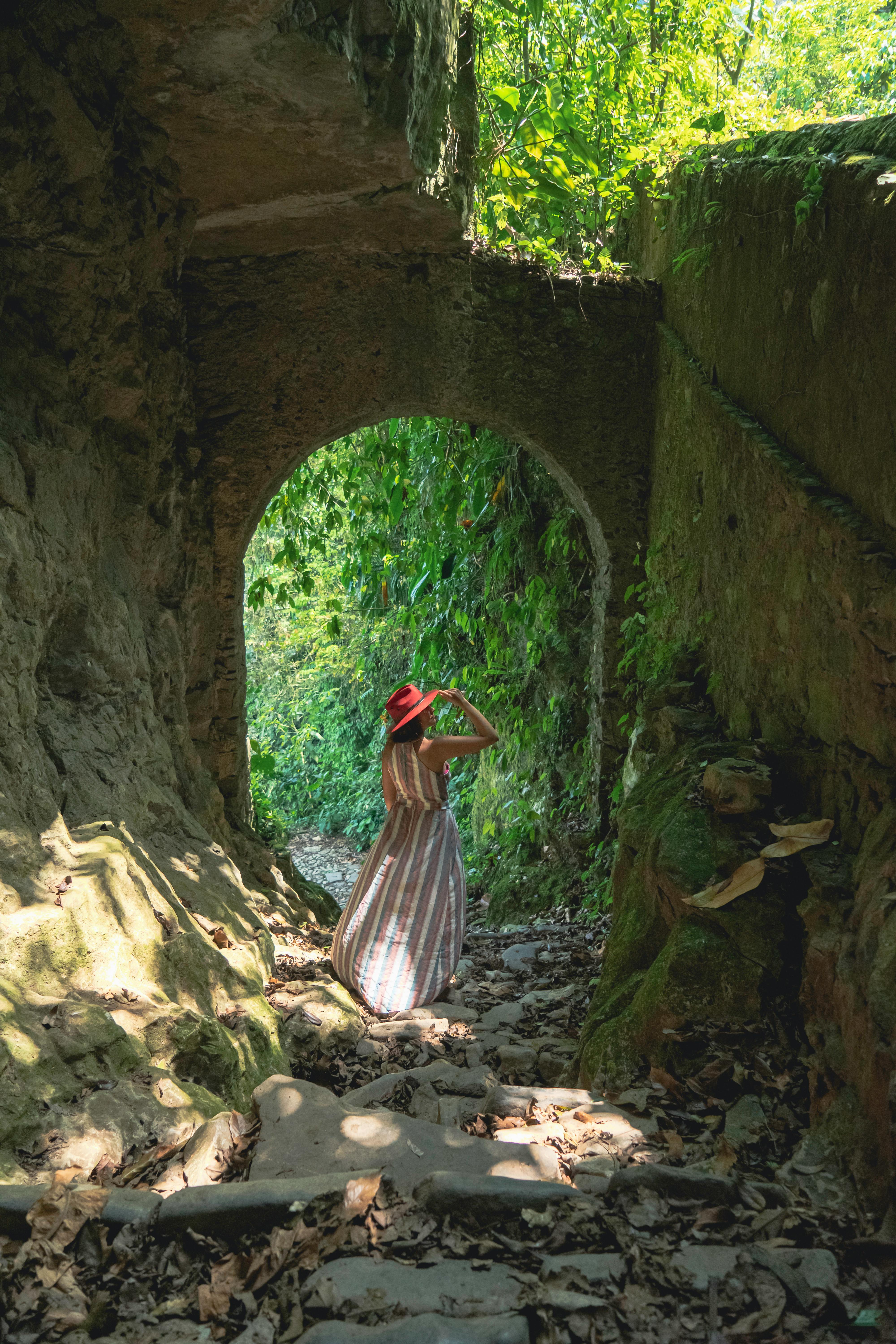 Free A woman in a red hat walks through a lush, overgrown stone tunnel in Cuetzalan, Mexico. Stock Photo