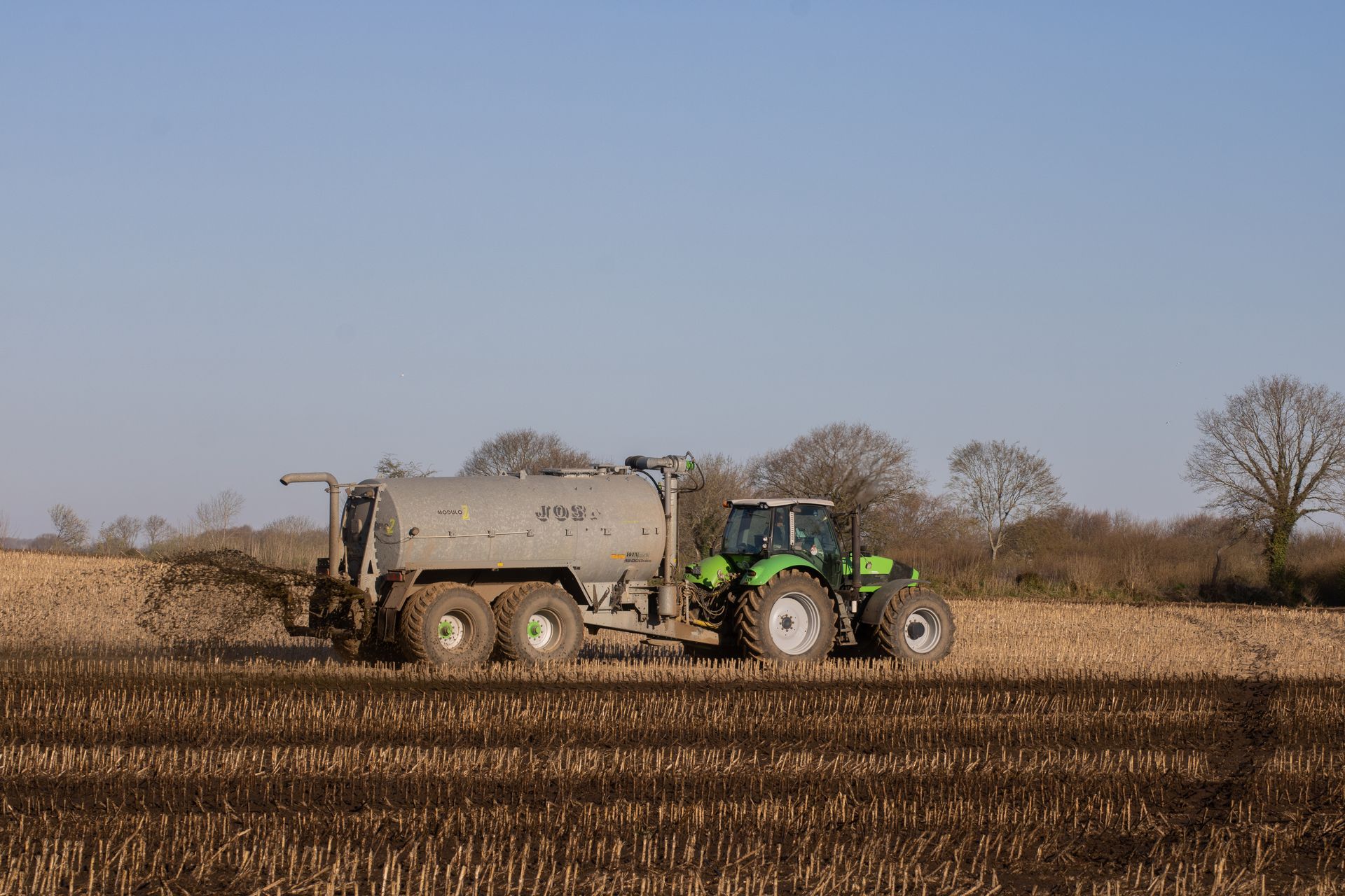 A green tractor spreading fertilizer on a brown, freshly plowed farmland under a clear sky.