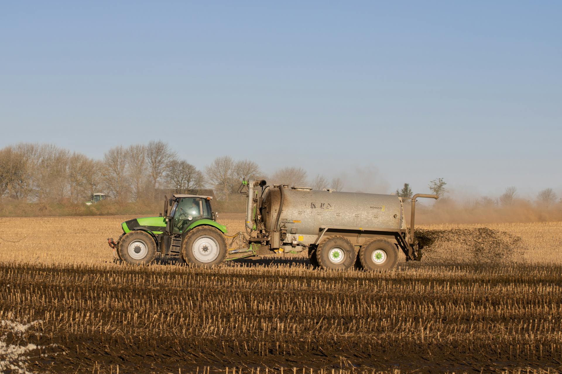 A tractor with a tanker spreading fertilizer over a barren farmland under clear skies.