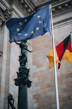 A striking view of the EU and German flags beside a historic sculpture at a landmark building.