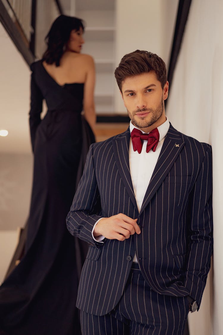 Man Posing In A Navy Blue Suit With Stripes And A Red Bow Tie And Woman Walking In A Long Black Dress