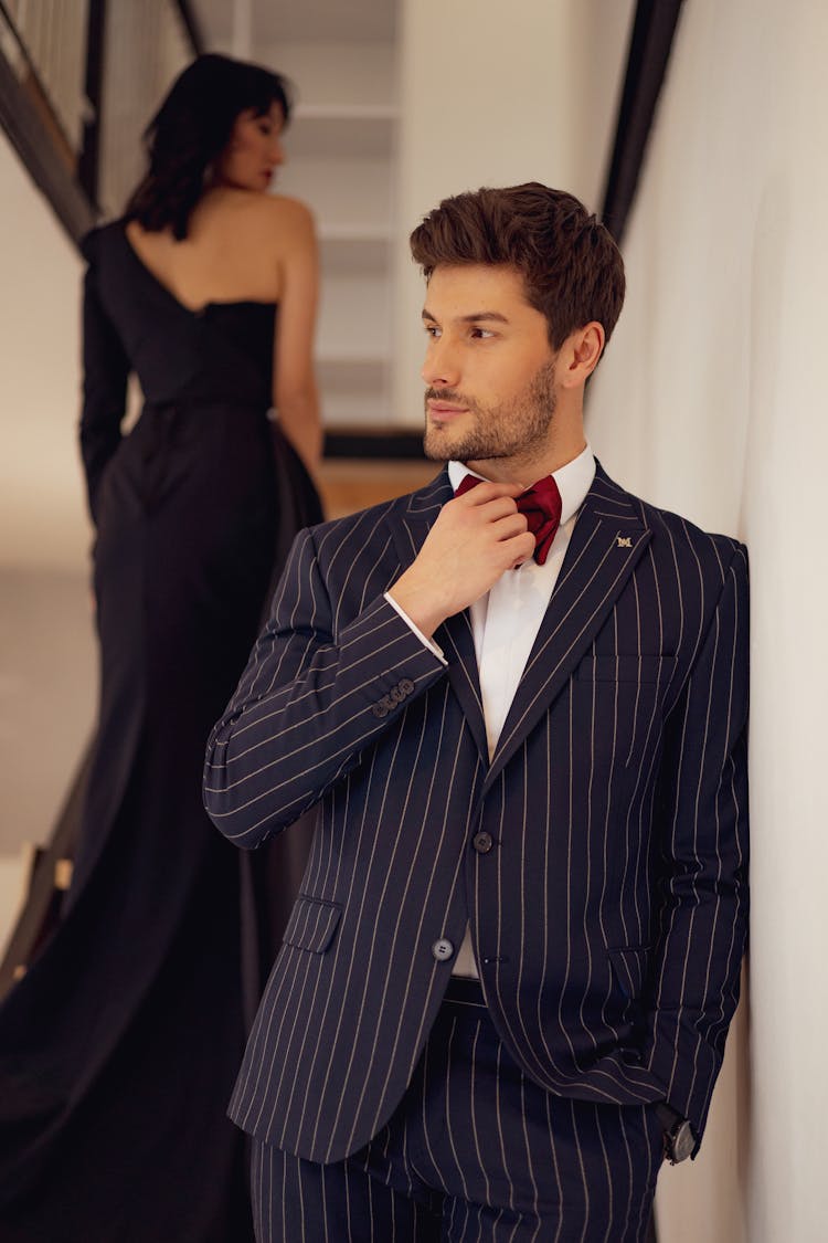 Man Posing In A Navy Blue Suit With Stripes And A Red Bow Tie And Woman Walking In A Long Black Dress