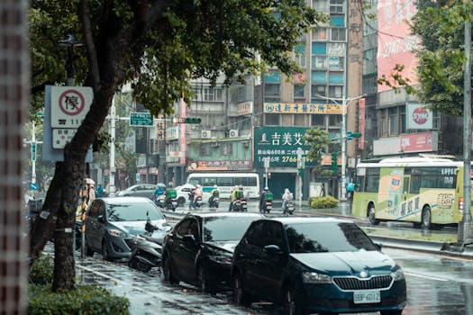 A bustling city street in the rain, featuring parked cars, buses, and motorbikes.