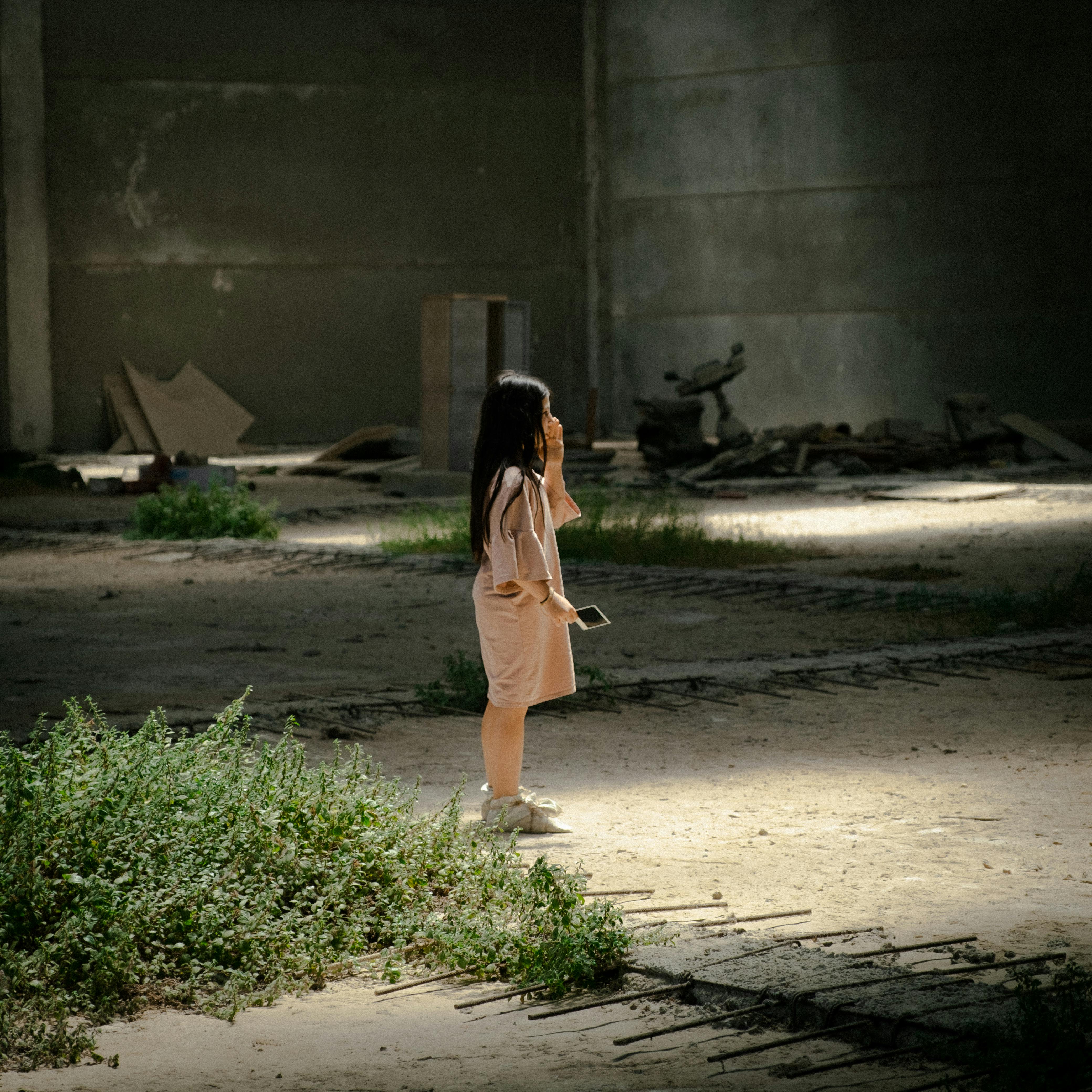 A young girl stands alone in an abandoned warehouse, suggesting introspection.